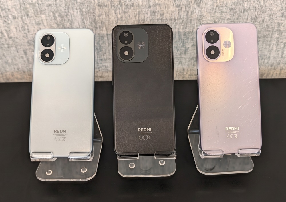 Three Redmi smartphones on display stands, back views in light blue, black, and lavender-pink, showing dual-camera modules and Redmi logo.
