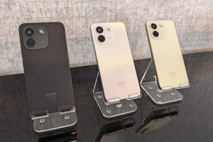Three Redmi smartphones on clear display stands, shown from the back in black, pearl gradient, and pale green colors.