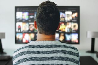 Person viewed from behind, wearing a striped sweater, looking at a wall-mounted screen with app thumbnails (streaming UI).