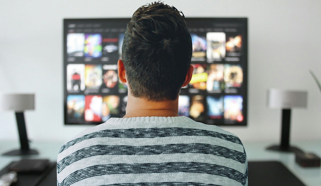 Person viewed from behind, wearing a striped sweater, looking at a wall-mounted screen with app thumbnails (streaming UI).