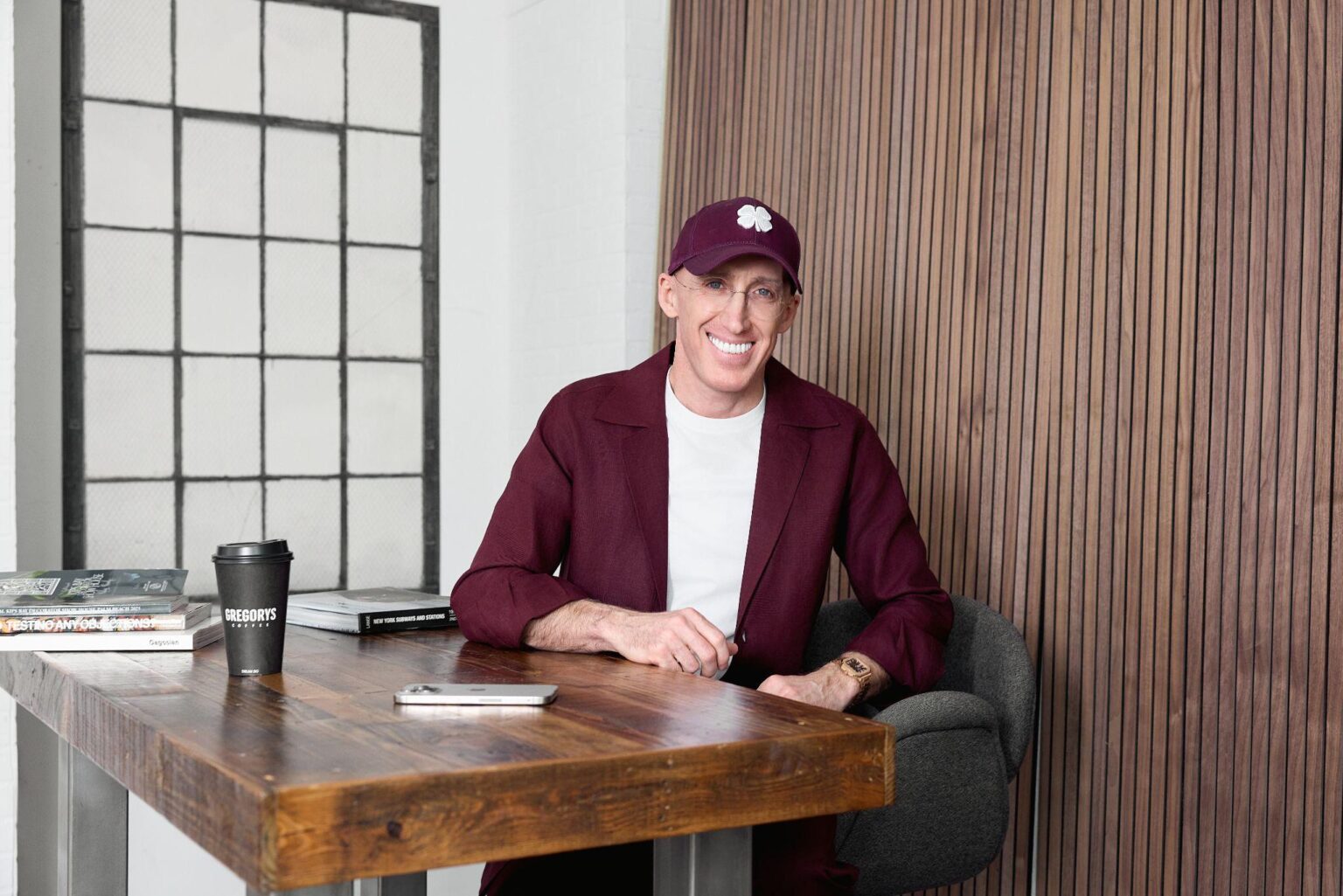 Smiling man in a maroon blazer and cap with a shamrock sits at a wooden table in a modern, brick-walled space with magazines and a coffee cup nearby.