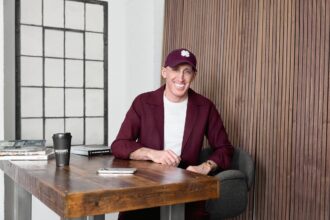 Smiling man in a maroon blazer and cap with a shamrock sits at a wooden table in a modern, brick-walled space with magazines and a coffee cup nearby.