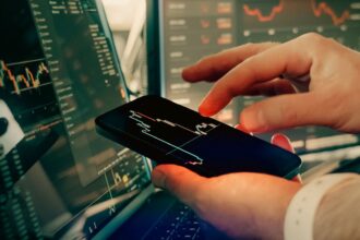 Person holding a smartphone displaying a stock chart while multiple monitors show market data in the background.