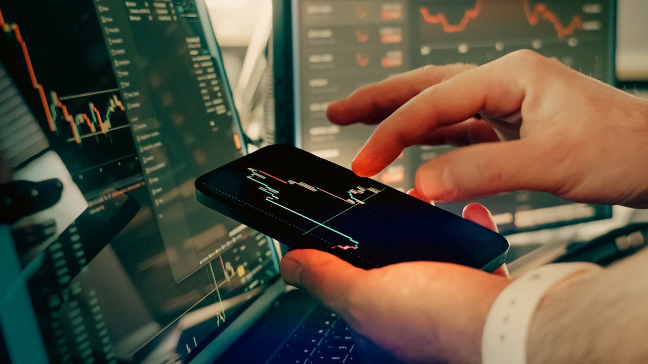 Person holding a smartphone displaying a stock chart while multiple monitors show market data in the background.