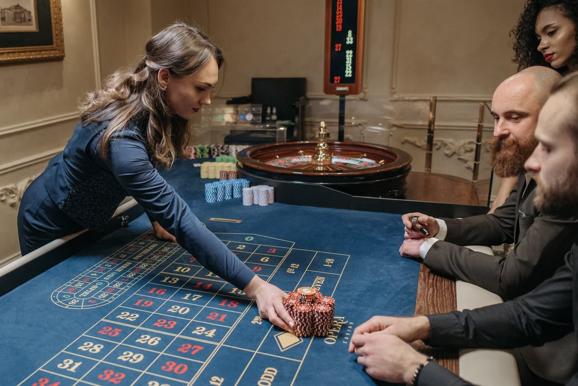 Female dealer arranges chips on a roulette table as two players watch intently, in a casino setting.