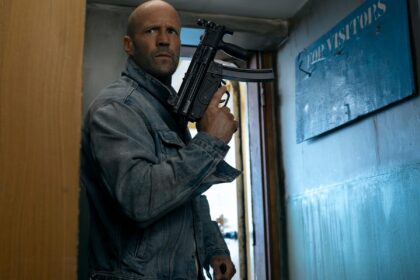 Man in a denim jacket stands in a narrow hallway, gripping a black rifle and looking alert.