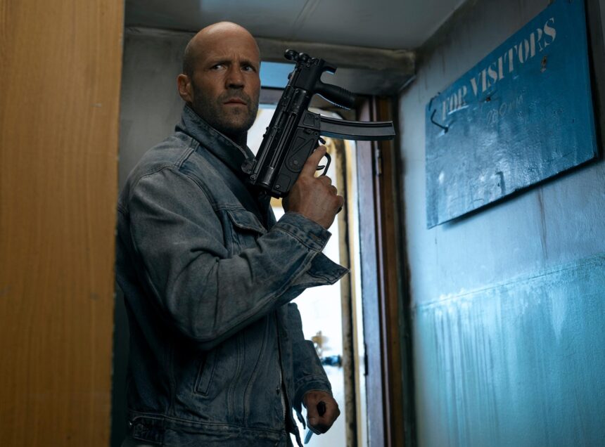 Man in a denim jacket stands in a narrow hallway, gripping a black rifle and looking alert.