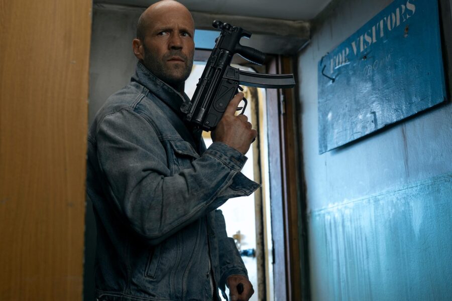 Man in a denim jacket stands in a narrow hallway, gripping a black rifle and looking alert.