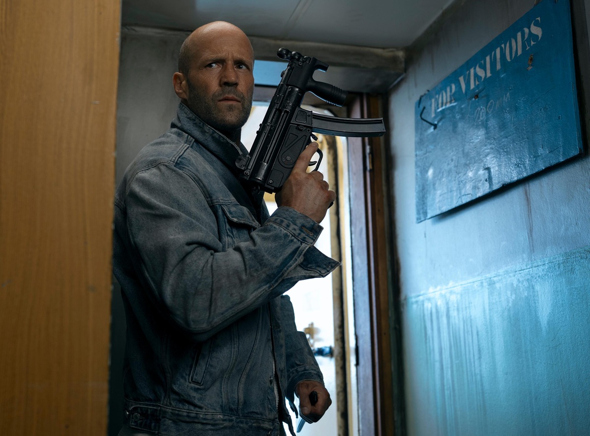 Man in a denim jacket stands in a narrow hallway, gripping a black rifle and looking alert.