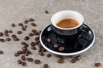 Black ceramic espresso cup on a matching saucer with scattered roasted coffee beans on a gray concrete surface.