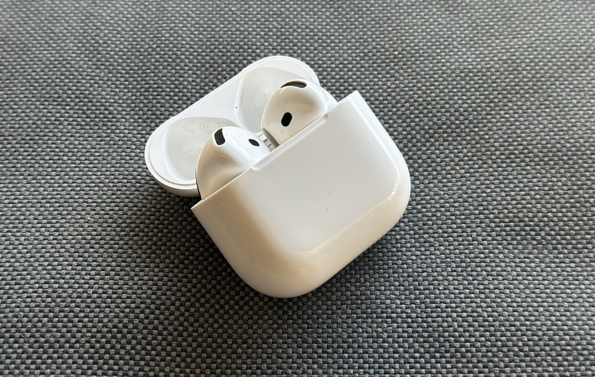 White wireless earbuds in an open charging case on a textured gray fabric surface.
