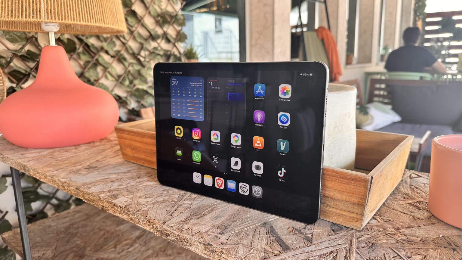 Tablet on a wooden table outdoors displaying app icons on the home screen (cafe-style setting).','Close-up of a tablet showing apps like App Store, Photos, Instagram on a patio table.','Tablet resting on a rustic wooden table with a pink lamp beside it, outdoor seating in the background.','Outdoorsy workspace: tablet with colorful app icons on a wood table and a woven lampshade nearby.','A tablet on a weathered wooden table beside a pink lamp, with blurred seating in the background.