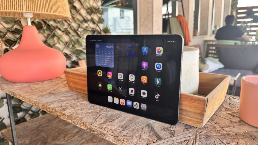 Tablet on a wooden table outdoors displaying app icons on the home screen (cafe-style setting).','Close-up of a tablet showing apps like App Store, Photos, Instagram on a patio table.','Tablet resting on a rustic wooden table with a pink lamp beside it, outdoor seating in the background.','Outdoorsy workspace: tablet with colorful app icons on a wood table and a woven lampshade nearby.','A tablet on a weathered wooden table beside a pink lamp, with blurred seating in the background.