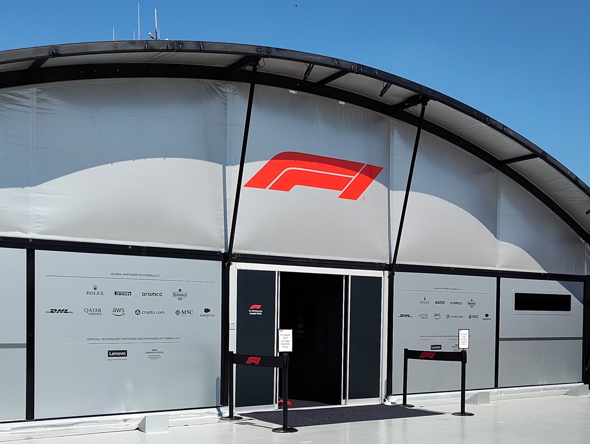 Formula 1 team hospitality tent with a large red F1 logo, open entrance, and sponsor banners on the side walls under a blue sky.