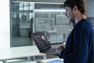 Engineer standing at a white desk, typing on a laptop displaying code in a modern office.