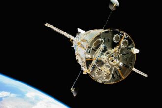 Spacecraft in orbit with a round hull and extended truss, set against the blackness of space and Earth's curved horizon below.