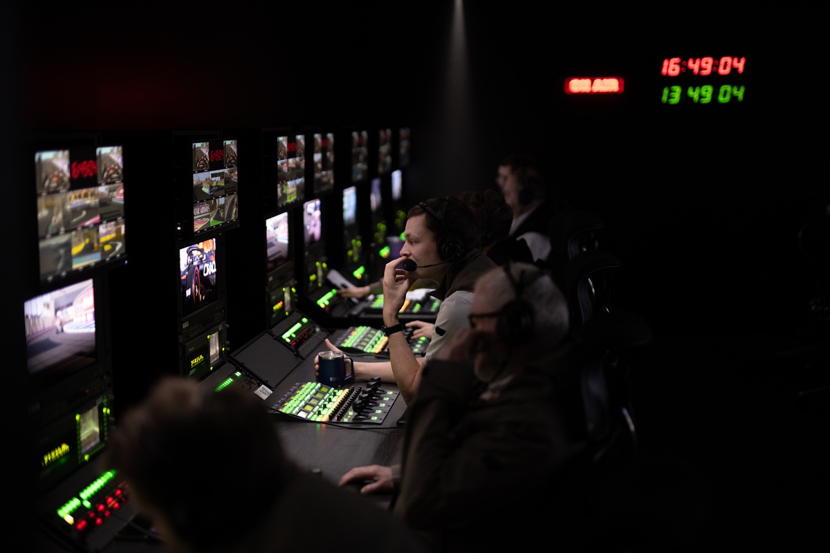 Broadcast control room with operators wearing headsets at a bank of monitors displaying game footage, illuminated by green and red control panels.