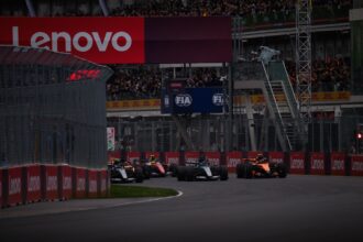 Formula 1 cars race down a track with red Lenovo banners along the barriers and a packed grandstand above the pit wall at dusk/overcast lighting.