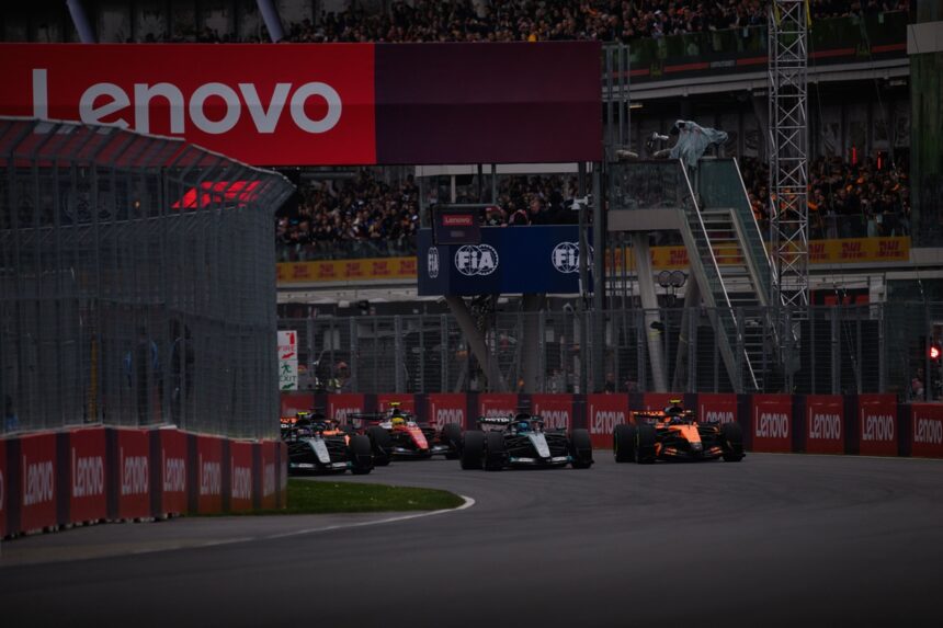 Formula 1 cars race down a track with red Lenovo banners along the barriers and a packed grandstand above the pit wall at dusk/overcast lighting.