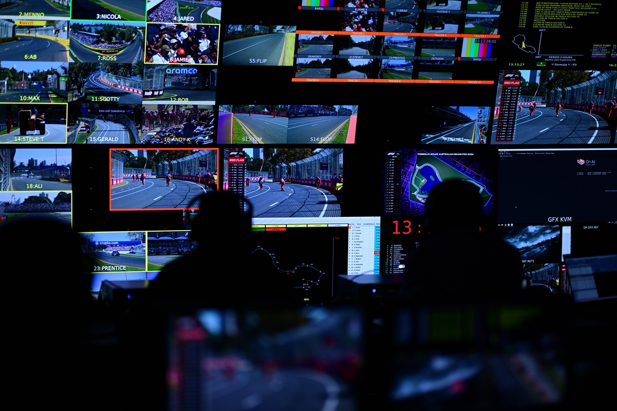 Control room with multiple wall-mounted screens showing racing feeds and timing data, silhouetted operators monitor the feeds