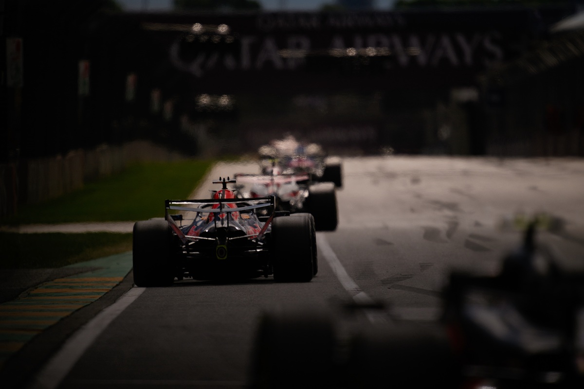 Rear view of a red Formula 1 car leading a line of race cars on a dimly lit track, highlighting wide tires and aero setup.