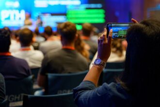 Person in the foreground holds up a smartphone to record a presentation at a conference, with attendees and large screens in the background