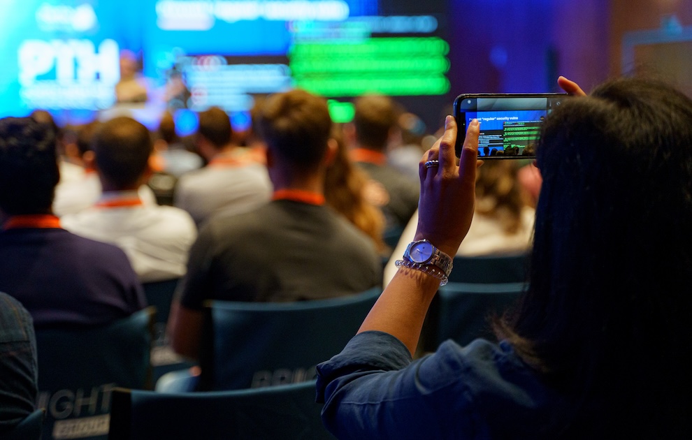 Person in the foreground holds up a smartphone to record a presentation at a conference, with attendees and large screens in the background