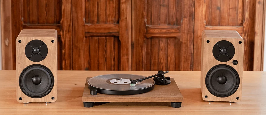 Turntable on a wooden table flanked by two matching wooden bookshelf speakers against a wood-paneled wall, with a vinyl record on the platter.