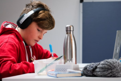 Teen student wearing a red hoodie and headphones writes in a notebook at a desk.