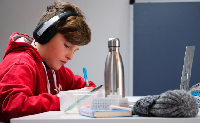 Teen student wearing a red hoodie and headphones writes in a notebook at a desk.