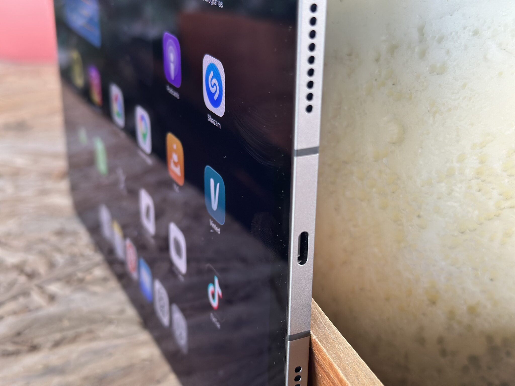 Close-up of a smartphone screen showing app icons (including Shazam) from a side angle, with a textured wall and wooden surface in the background.