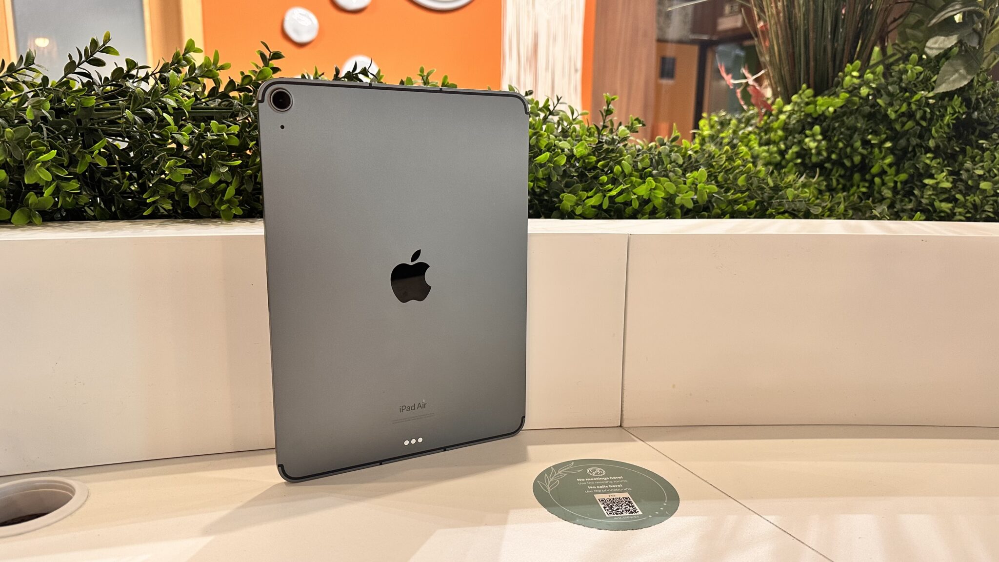 Back of a silver iPad Air stood upright on a white display counter with green bushes and an orange wall in the background.