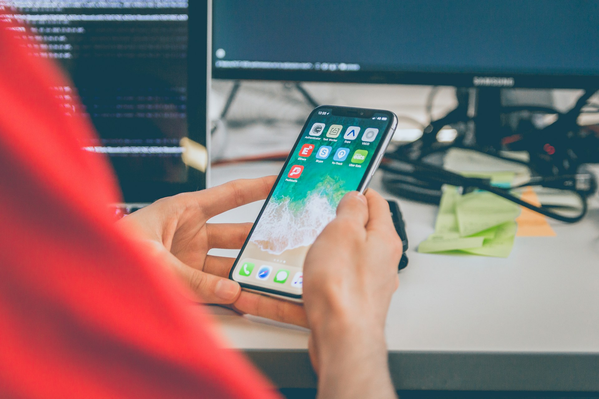 Person holding a smartphone showing app icons on a desk with a computer monitor nearby and sticky notes nearby.