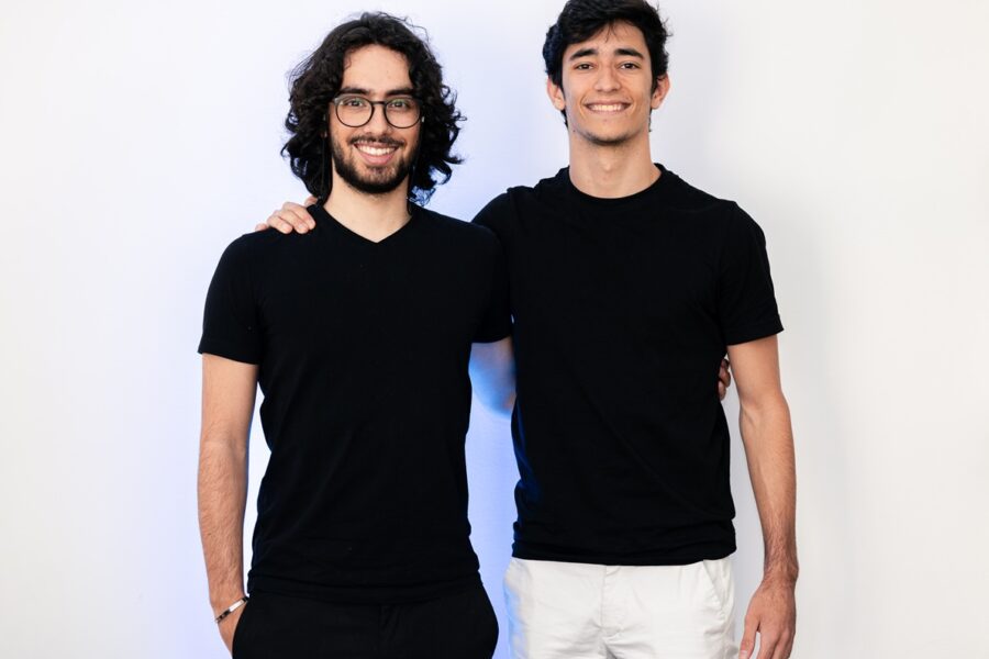 Two men in black T-shirts stand side by side, one with glasses, smiling at the camera with an arm around the other's shoulder.