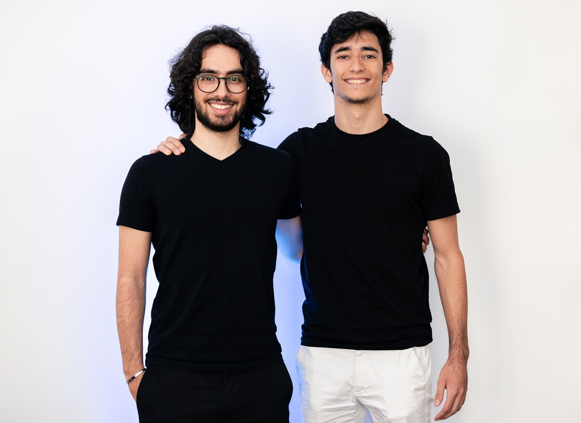 Two men in black T-shirts stand side by side, one with glasses, smiling at the camera with an arm around the other's shoulder.