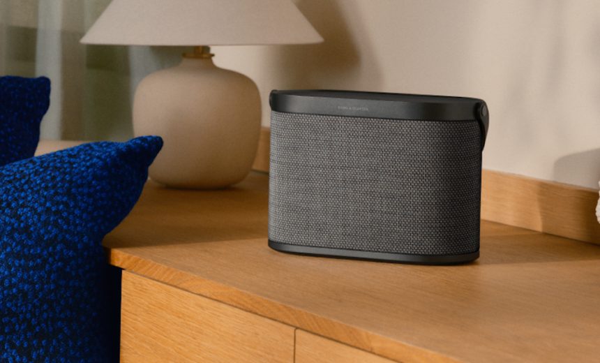 Portable Bluetooth speaker on a wooden dresser with a beige lamp in the background and blue cushions nearby.
