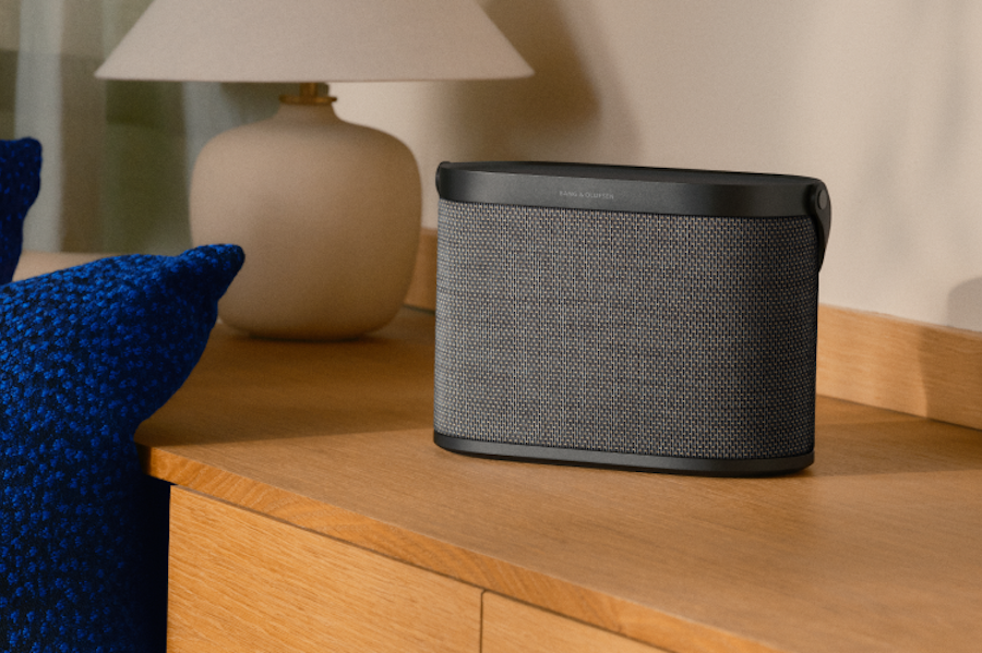 Portable Bluetooth speaker on a wooden dresser with a beige lamp in the background and blue cushions nearby.