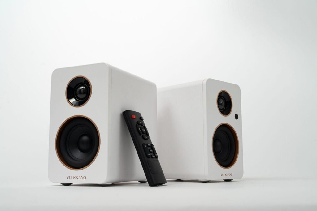Pair of white bookshelf speakers with black drivers and a remote control resting between them, on a white surface (VULKKANO).