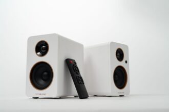 Pair of white bookshelf speakers with black drivers and a remote control resting between them, on a white surface (VULKKANO).