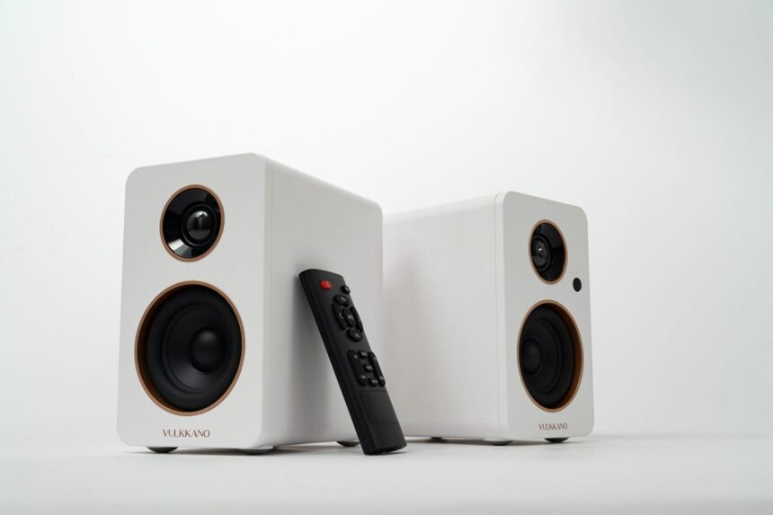 Pair of white bookshelf speakers with black drivers and a remote control resting between them, on a white surface (VULKKANO).