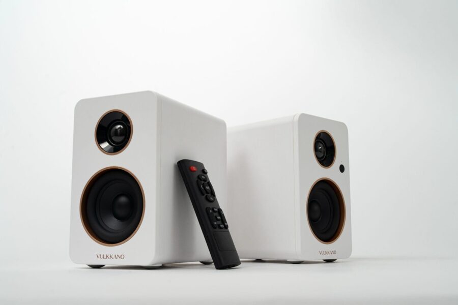 Pair of white bookshelf speakers with black drivers and a remote control resting between them, on a white surface (VULKKANO).
