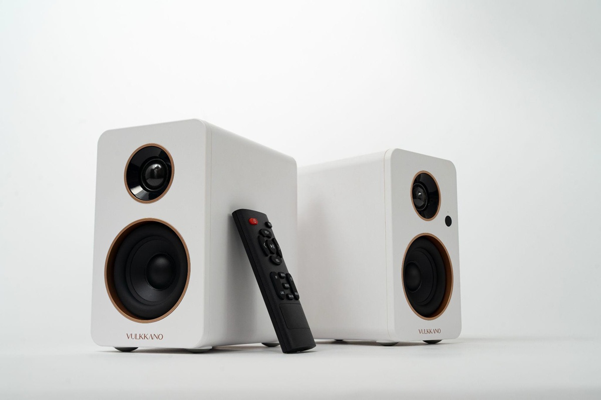 Pair of white bookshelf speakers with black drivers and a remote control resting between them, on a white surface (VULKKANO).