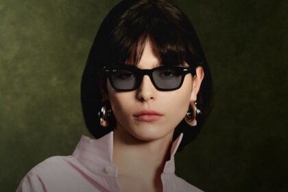 Woman with short dark hair wearing black sunglasses and large hoop earrings, facing the camera against a dark green background.