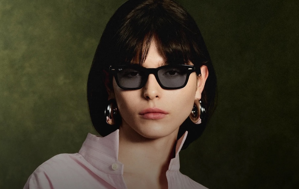 Woman with short dark hair wearing black sunglasses and large hoop earrings, facing the camera against a dark green background.