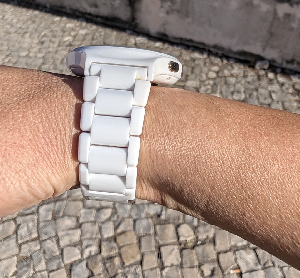 Close-up of a white ceramic wristwatch on a tan wrist against a cobblestone pavement outside.