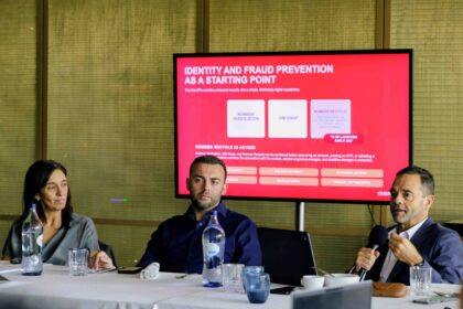 Panel of three adults seated at a conference table with water bottles and glasses, while a red presentation slide is projected on a screen behind them.