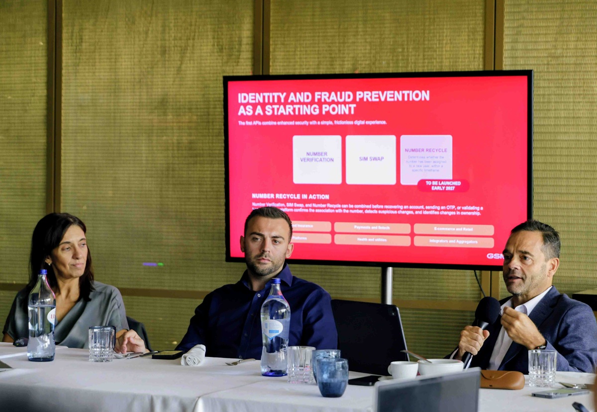 Panel of three adults seated at a conference table with water bottles and glasses, while a red presentation slide is projected on a screen behind them.