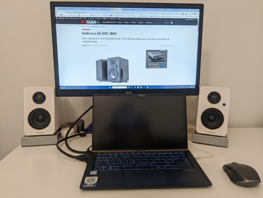 Desk setup with a laptop in front of a larger monitor, flanked by two white speakers on stands and a wireless mouse on the right.