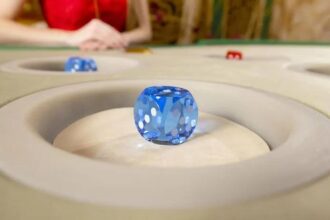 Close-up of a blue translucent die resting in a recessed circular area of a tabletop game, with a blurred player in red in the background.