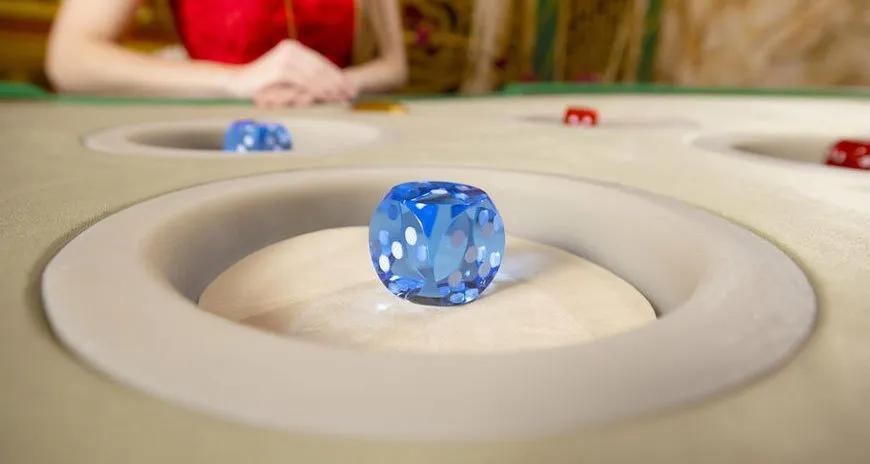 Close-up of a blue translucent die resting in a recessed circular area of a tabletop game, with a blurred player in red in the background.